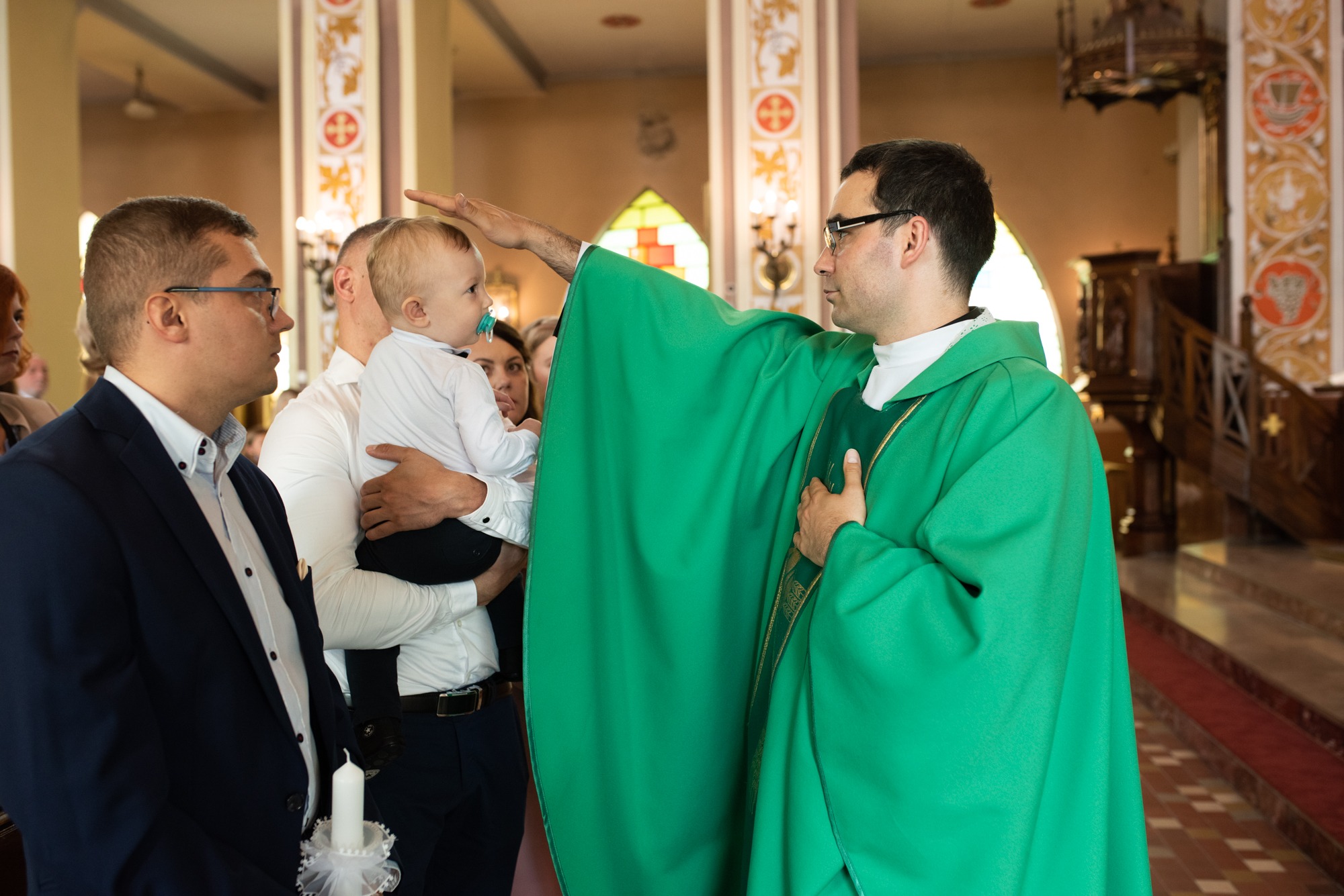Ksiądz w zielonej szacie udziela błogosławieństwa niemowlęciu na rękach ojca w kościele, obok mężczyzna trzyma świecę.