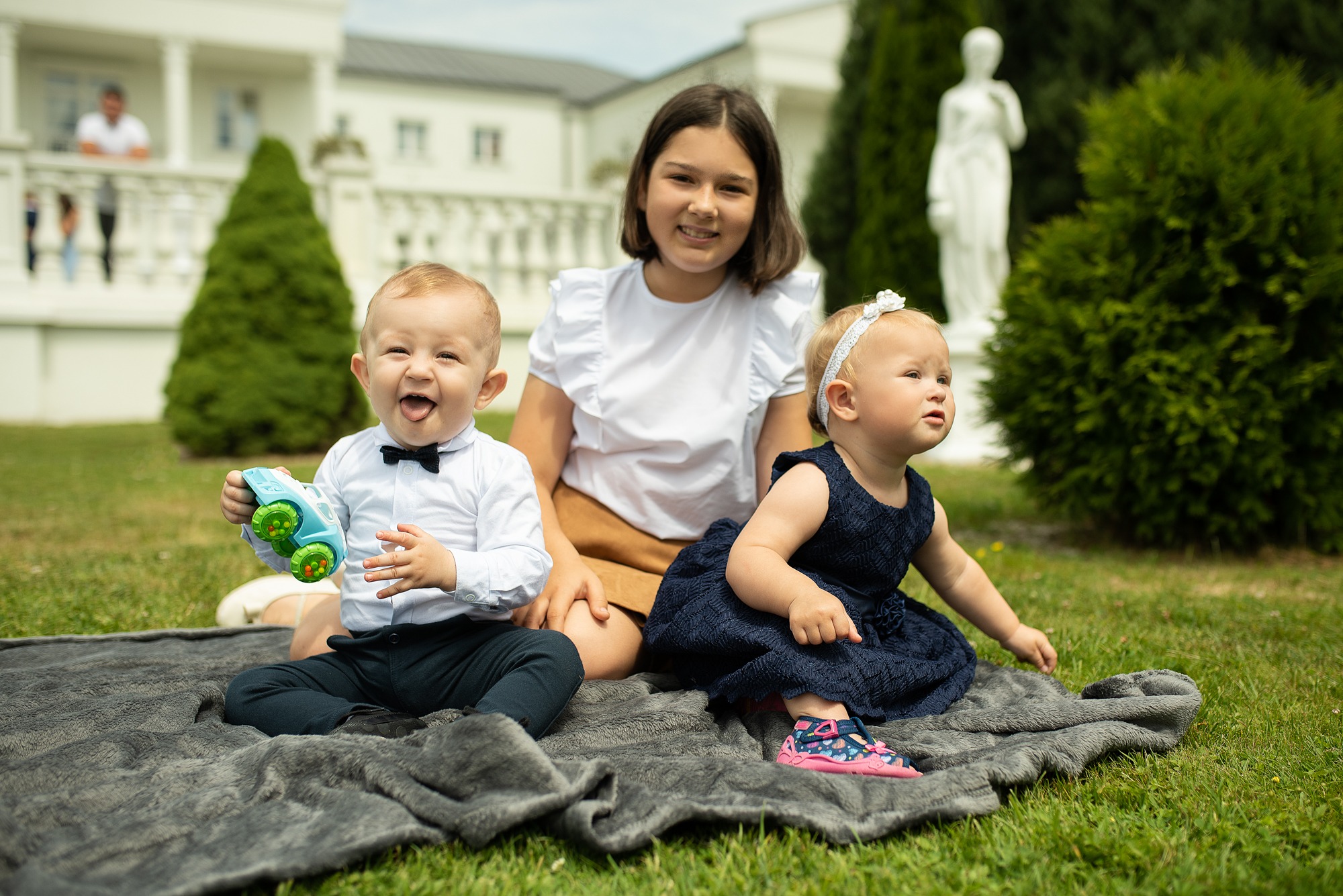 Rodzinne zdjęcie na trawie: niemowlak z zabawką, dziewczynka w sukience z opaską i nastolatka w tle, biała rezydencja i statua w ogrodzie.