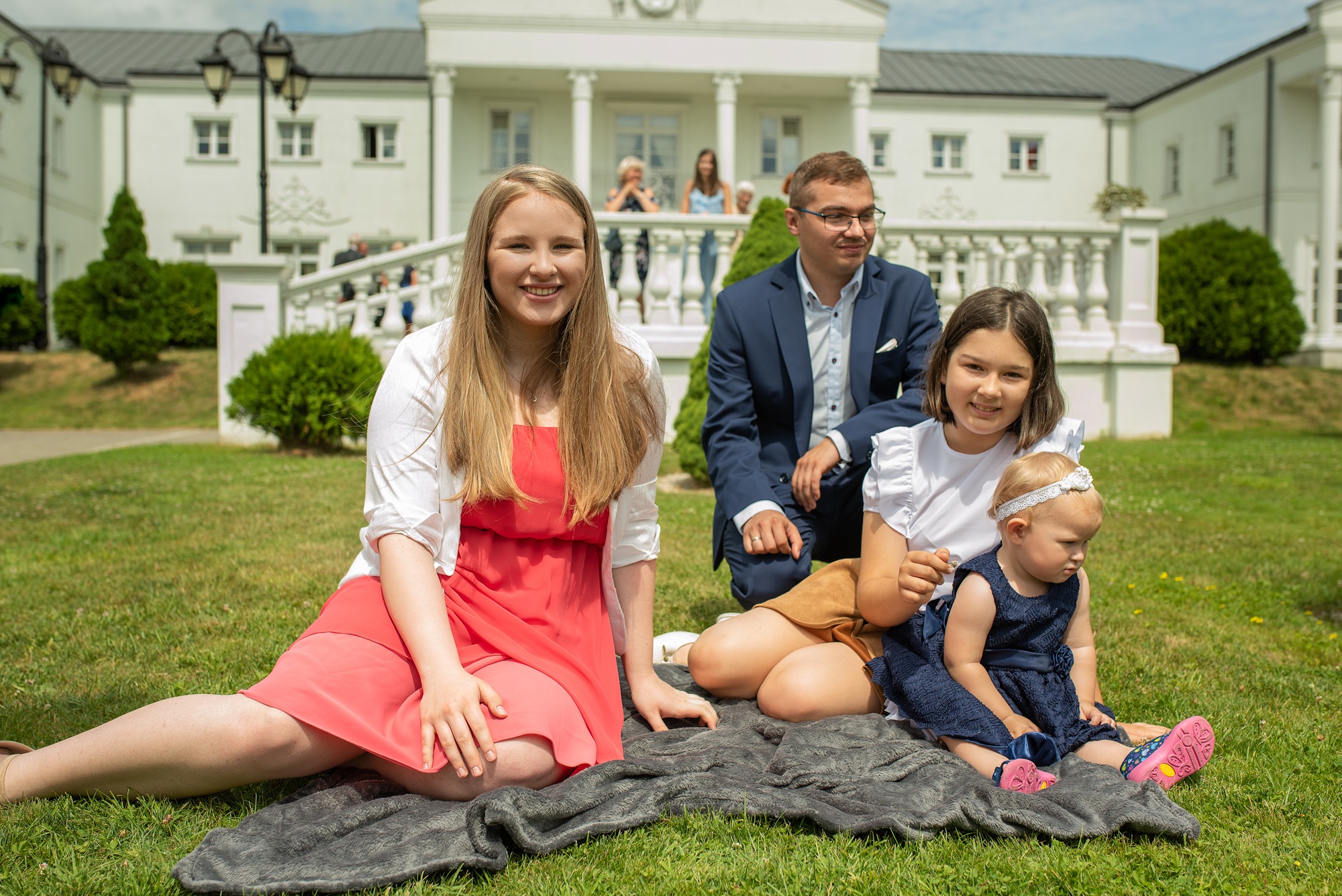 Rodzina pozuje na trawie przed białym budynkiem z kolumnami, matka w koralowej sukience, ojciec w granatowym garniturze, córki w bieli i granacie, siedzą na szarym kocu.