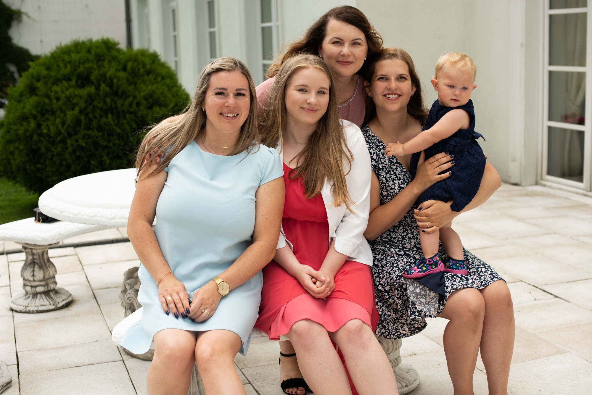 Portret rodzinny: cztery kobiety i małe dziecko w sukienkach, siedzące na kamiennej ławce na tle białego budynku i zielonego krzewu.