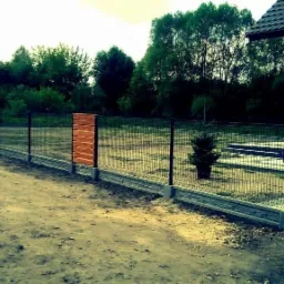 Nowoczesne ogrodzenie panelowe z drewnianymi akcentami, betonowa podmurówka z dekoracyjnym wzorem, brama z poziomych desek, mała choinka w doniczce wewnątrz posesji.