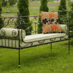 Kuta metalowa ławka ogrodowa z zielonymi poduszkami i poduszką dekoracyjną w kwiaty, stojąca na trawie przed iglakami.