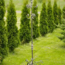 Kuty, metalowy wieszak na ubrania stojący na trawniku przed rzędem tuj, z widocznymi zdobieniami w formie spiral.