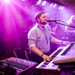 Zespół muzyczny grający na żywo na scenie oświetlonej fioletowymi światłami, z widocznymi saksofonami w tle i mikserem Tascam na pierwszym planie.