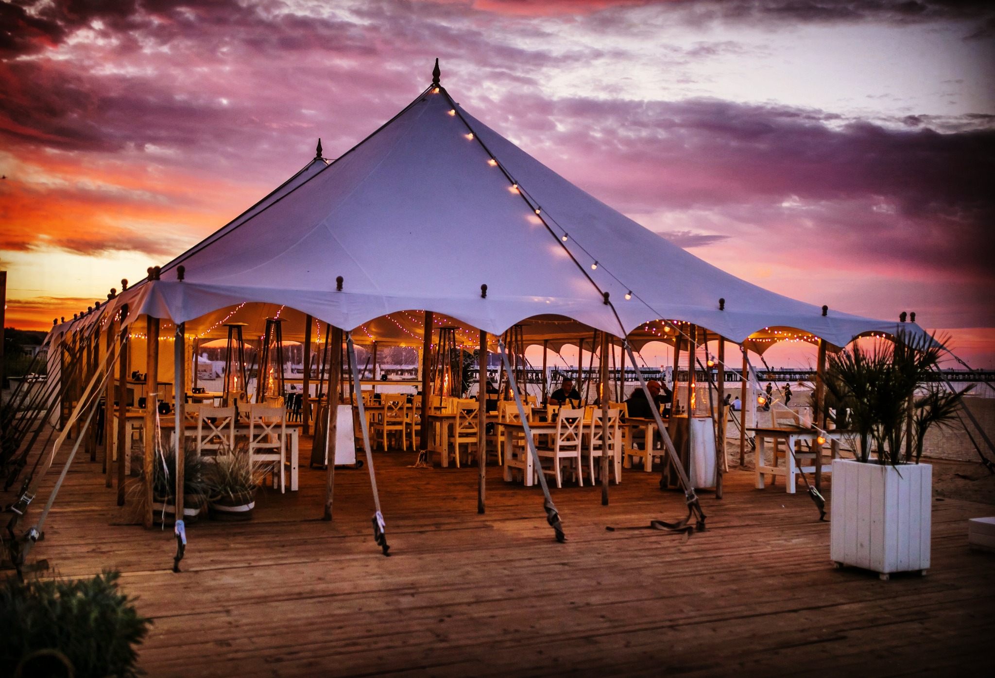 Rozświetlony namiot imprezowy na drewnianym podeście, z widocznymi stolikami i krzesłami, oświetlony girlandami żarówek, na tle zachodzącego słońca z purpurowymi chmurami.