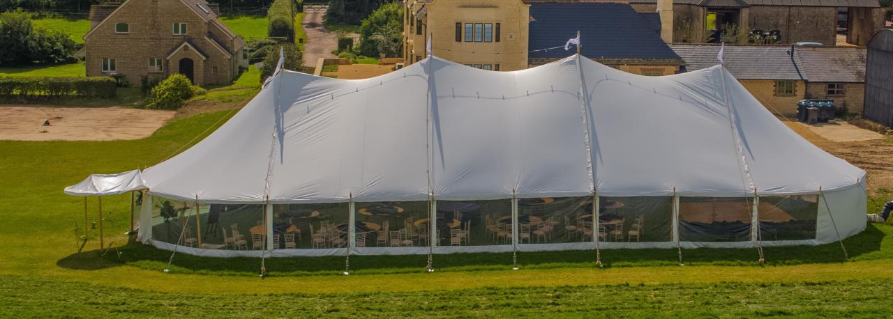 Rozległy biały namiot imprezowy na zielonej trawie, z widocznymi stołami i krzesłami wewnątrz, w tle budynki z kamienia