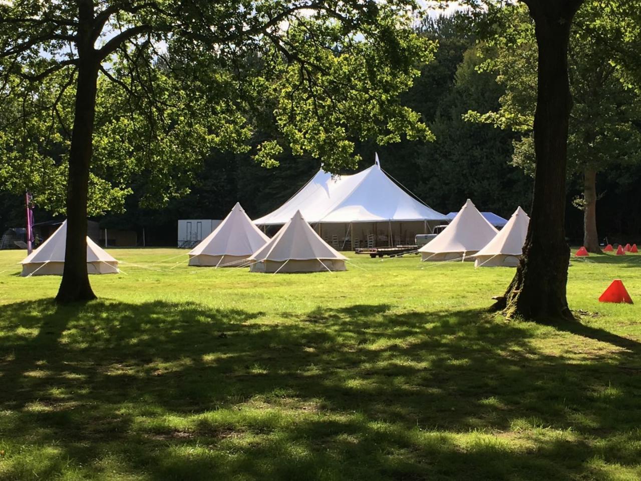 Rozległy teren zielony z rozstawionymi białymi namiotami tipi i dużym namiotem imprezowym w tle, oświetlony przez promienie słoneczne przebijające się przez korony drzew.