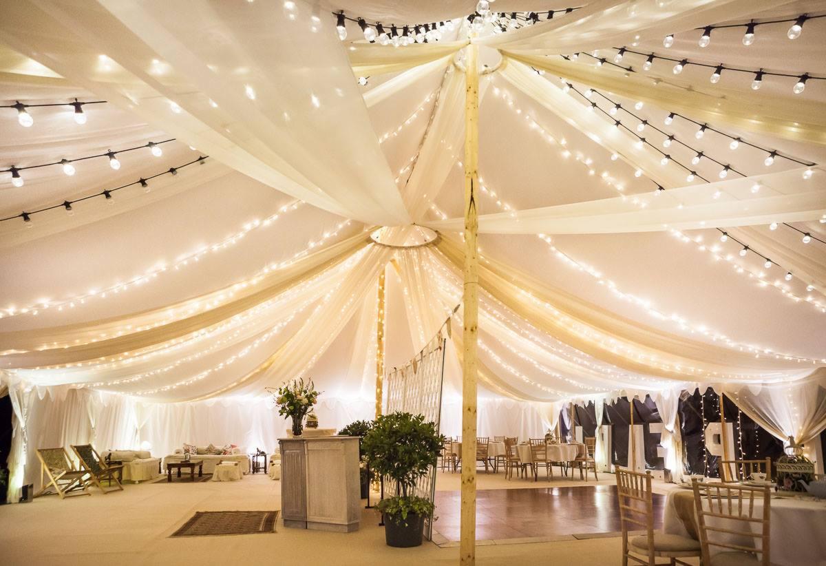Uroczysty namiot z beżowymi draperiami, oświetlony girlandami z żarówek, z barem, roślinami w donicach i miejscami do siedzenia.