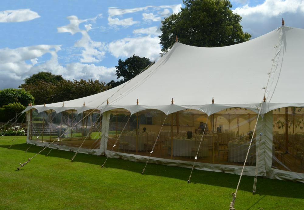 Duży, biały namiot imprezowy z przezroczystymi ścianami bocznymi, ustawiony na zielonym trawniku pod błękitnym niebem z chmurami. Wewnątrz widoczne stoły i krzesła przygotowane na uroczystość.
