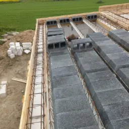 Wznoszenie ścian z szarych bloczków betonowych, zbrojenie stalowe w drewnianym szalunku. W tle zielone pole i budynek gospodarczy.