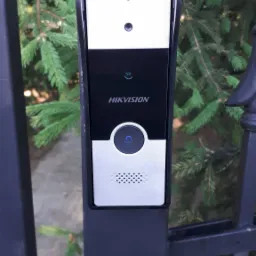 Zamontowany wideodomofon Hikvision na czarnym słupku bramy, z widocznym przyciskiem dzwonka i kamerą, na tle zielonych gałęzi drzewa iglastego.