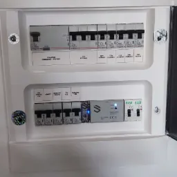 Rozdzielnica elektryczna z zabezpieczeniami różnicowoprądowymi i nadprądowymi firmy Legrand, zawierająca moduł sterowania inteligentnego domu z niebieską diodą LED, zamontowana w białej obudowie...