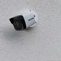 Kamera monitoringu Hikvision zamontowana na białej, chropowatej ścianie budynku, widok z dołu.