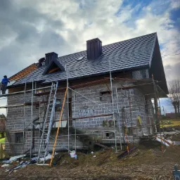 Montaż czarnej dachówki na nowym domu w trakcie budowy, widoczne rusztowania, drabina i pracownicy na dachu.