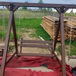 Drewniana huśtawka ogrodowa w kolorze ciemnego brązu, z siedziskiem dla dorosłych i małym siedziskiem dla dziecka, zawieszona na łańcuchach i linach, na tle sterty desek i ogrodzenia wiejskiego.