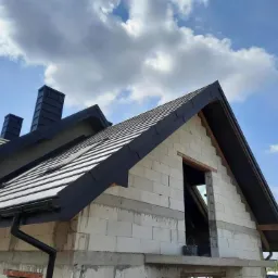 Dom w trakcie budowy z nowym, czarnym dachem z dachówki ceramicznej, widoczne kominy i rynny, ściany z białych bloczków, otwarty otwór okienny.