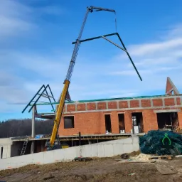 Żuraw montuje element konstrukcyjny dachu na budowie domu z czerwonej cegły, widoczna niezabezpieczona przestrzeń i sterta materiałów budowlanych w tle.