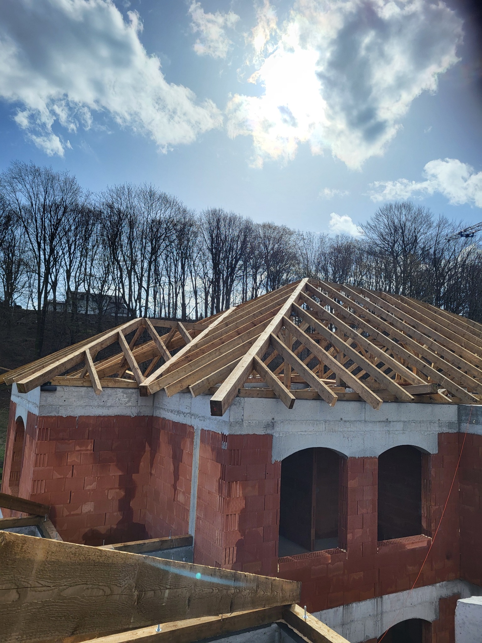Konstrukcja drewniana dachu w trakcie budowy, widoczne krokwie i murłat na budynku z czerwonej cegły, w tle drzewa i błękitne niebo z chmurami.