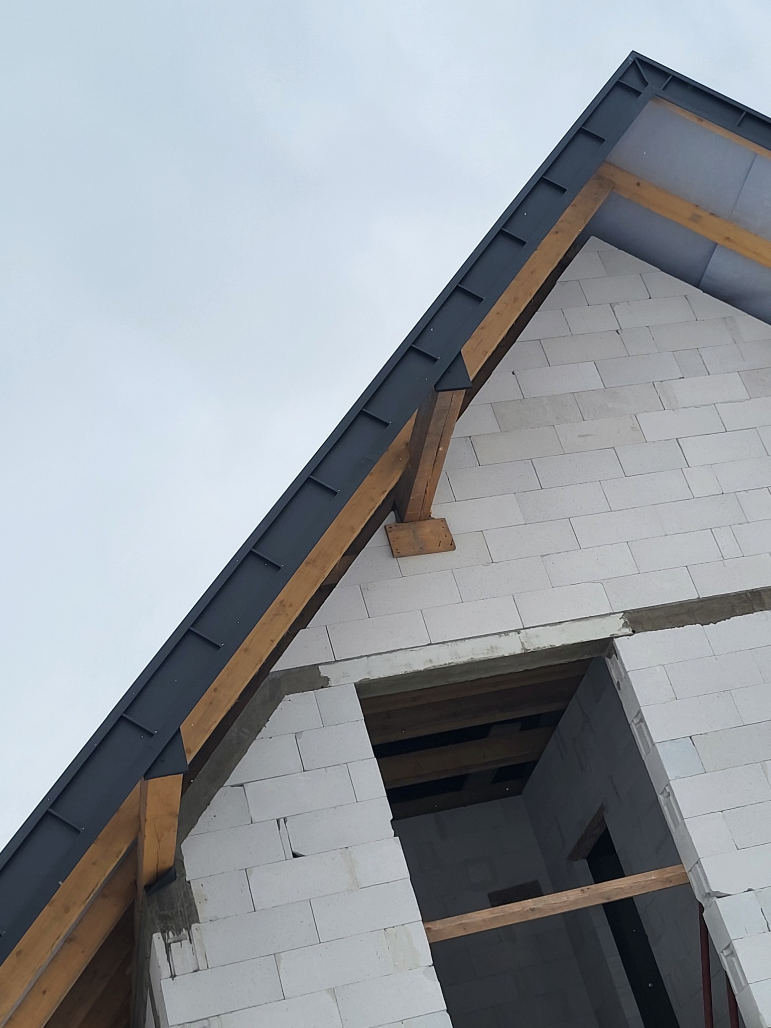 Fragment budynku w trakcie budowy: ściana z białych bloczków, widoczny otwór okienny, drewniana więźba dachowa i fragment ciemnej blachy na dachu, ujęcie z dołu pod kątem.