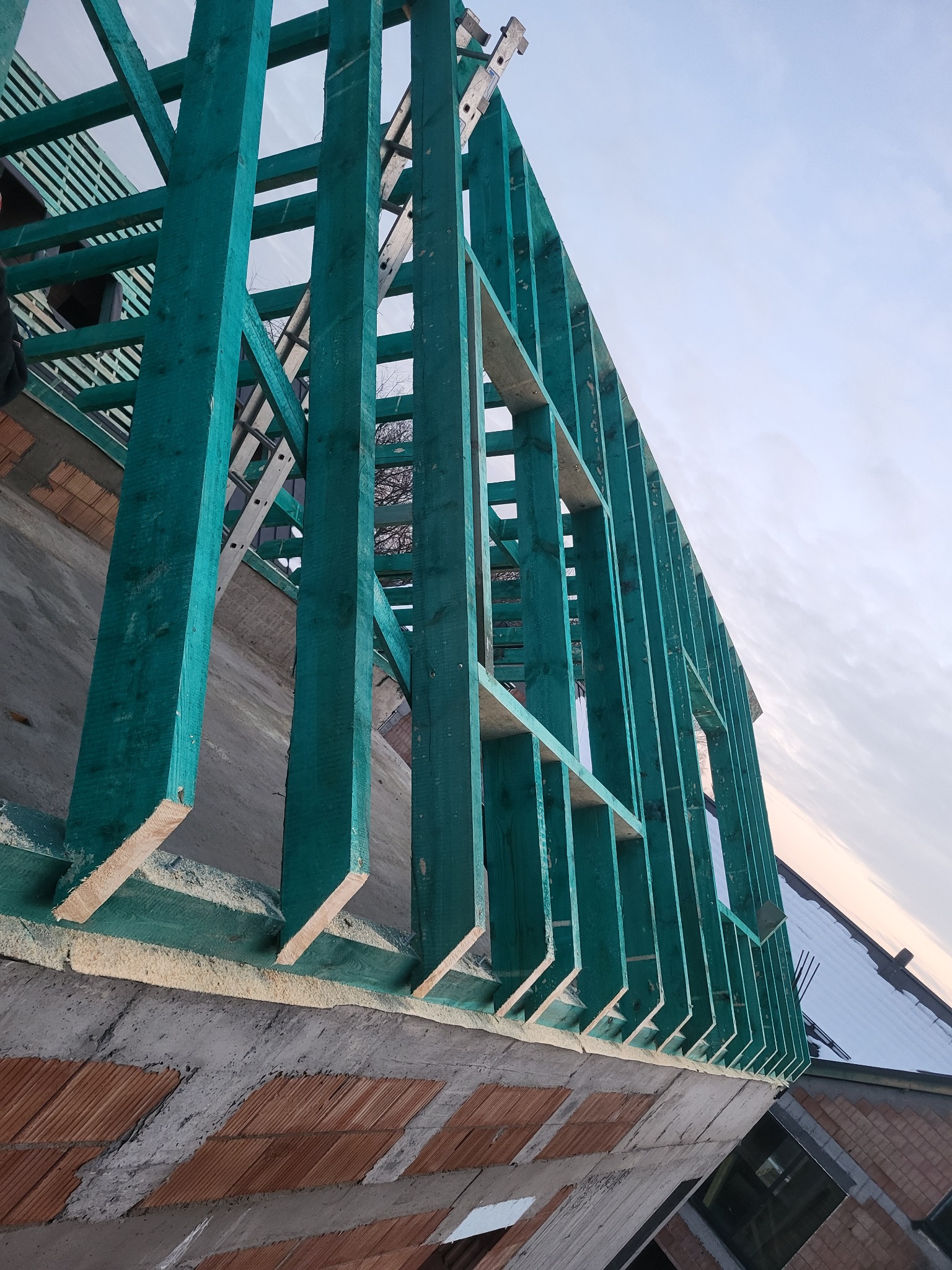 Fragment konstrukcji drewnianej, pomalowanej na zielono, w trakcie budowy dachu, widoczne cegły i drabina oparta o konstrukcję, jasne niebo w tle.