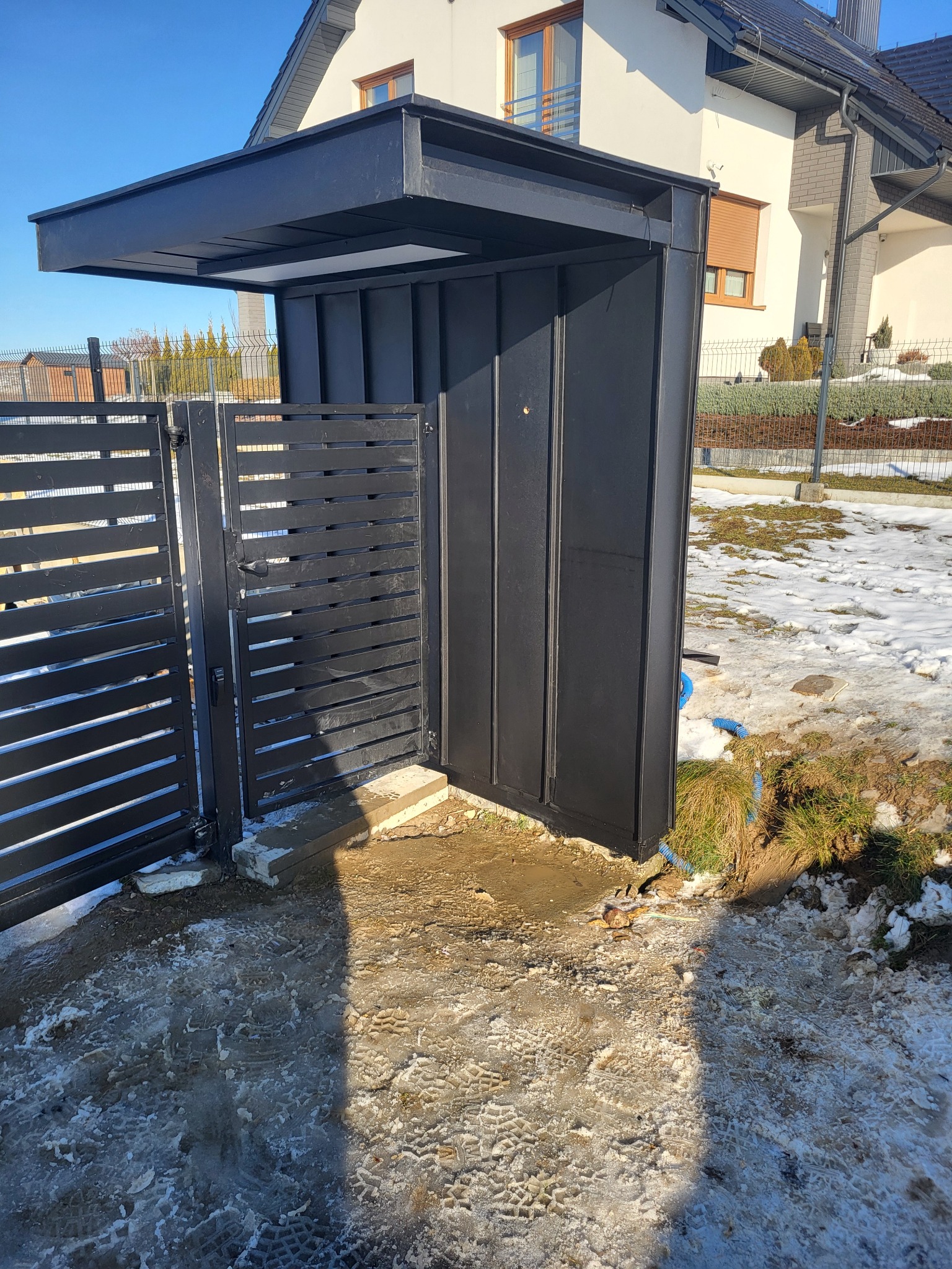 Nowoczesna, czarna wiata śmietnikowa z metalową bramą wjazdową na posesję, widoczna na tle domu jednorodzinnego w zimowej scenerii.