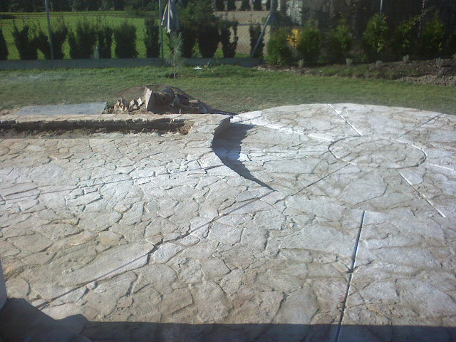 Niedokończona okrągła powierzchnia z nieregularnych kamieni w odcieniach szarości i beżu, z wyznaczonym wzorem koła i promienistych linii; w tle trawnik i krzewy.