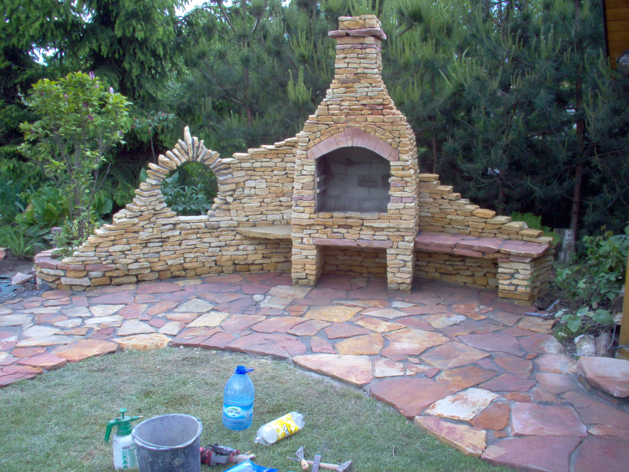 Kamienny grill ogrodowy z paleniskiem, łukiem i bocznymi siedziskami, otoczony ścieżką z nieregularnych płyt kamiennych, na trawniku widoczne narzędzia i butelki.