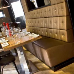 Brązowa tapicerowana ławka ze ścianką w restauracji, drewniany stół z metalową podstawą, nakryty do posiłku, czarne krzesła.