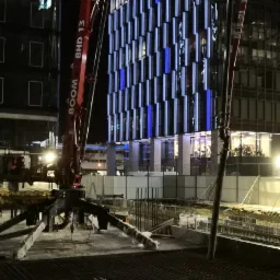 Nocna sceneria budowy w Gdańsku: pompa do betonu BOOM BHD 13 w akcji, nowoczesny budynek w tle z podświetlaną fasadą, widoczna siatka zbrojeniowa.