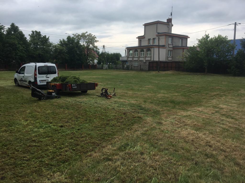 Trawa skoszona w Hajnówce, z przyczepką pełną zieleni, kosiarką Stiga i busem w tle, na rozległym trawniku przed budynkiem.