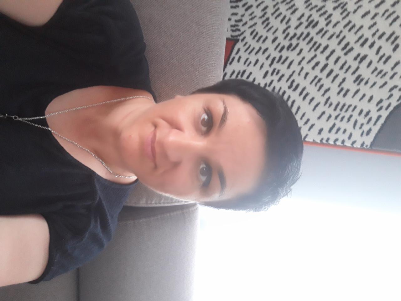 Portret kobiety z krótkimi, ciemnymi włosami, ubranej w ciemną koszulkę i srebrny łańcuszek, leżącej na szarej kanapie z wzorzystą poduszką w tle.