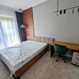 Nowoczesna sypialnia z łóżkiem zabezpieczonym folią, drewnianym biurkiem i designerską lampą sufitową na tle szarej ściany.