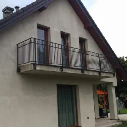 Czarna, metalowa balustrada balkonowa z pionowymi szczeblami, zamontowana przy elewacji domu z trzema oknami balkonowymi, widok z dołu na tle pochmurnego nieba.