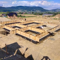 Usługi ogólnobudowlane - Szalunek fundamentów domu jednorodzinnego z hydroizolacją, widok na góry i zabudowę mieszkalną, na placu budowy pracownik w białym kasku i ciężarówka betoniarni.