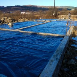 Usługi ogólnobudowlane - Zalane betonem ławy fundamentowe zabezpieczone niebieską folią, widoczna pompa do betonu, w tle pagórkowaty krajobraz.