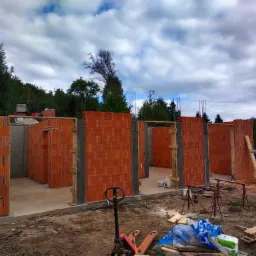 Wznoszenie ścian domu z czerwonej cegły. Widoczne betonowe słupy, drewniane szalunki i narzędzia. W tle zielone drzewa i pochmurne niebo.