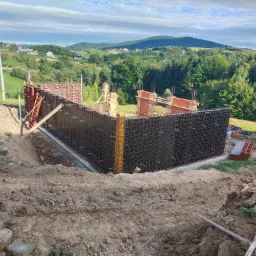 Zbrojenie fundamentów pod budowę domu na wzgórzu, widok na konstrukcję z prętów, szalunki i mini koparkę, malowniczy krajobraz w tle.