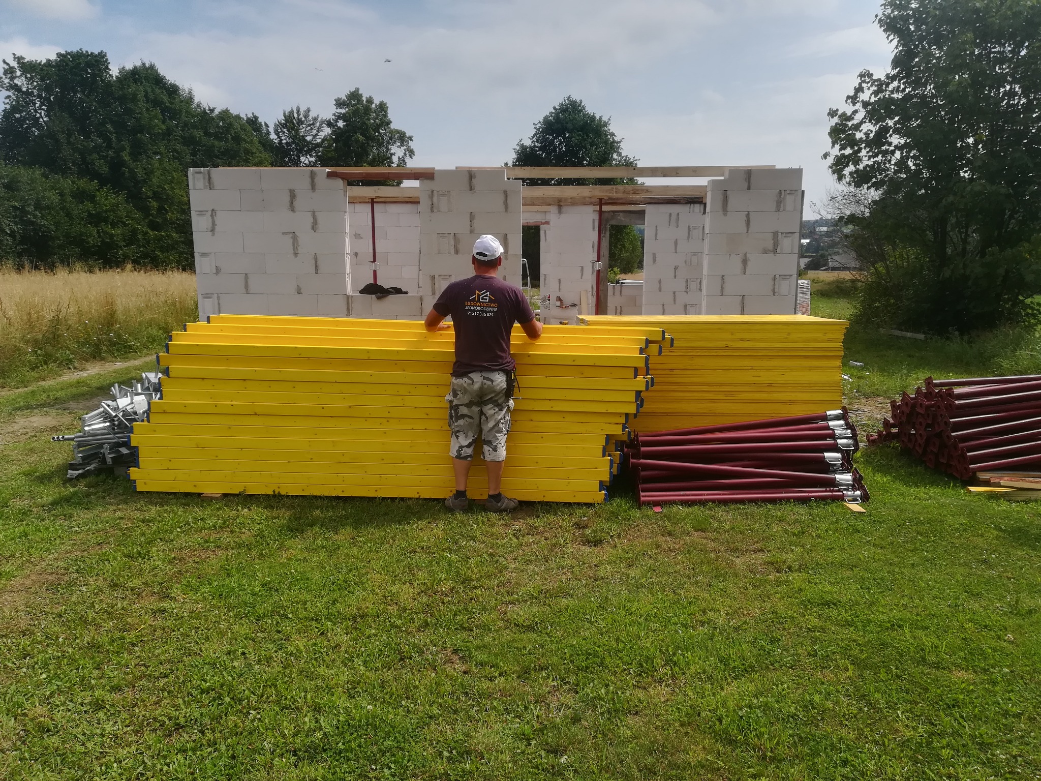 Budowa domu z betonu komórkowego: murarz układa żółte szalunki przed ścianą z pustaków, widoczne podpory szalunkowe i logo firmy na koszulce pracownika.