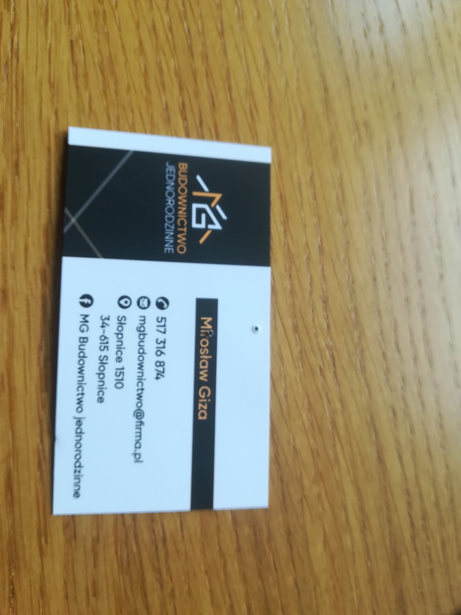 Wizytówka firmy budowlanej MG Budownictwo Jednorodzinne z danymi kontaktowymi Mrosława Gizy, leżąca na drewnianym blacie.