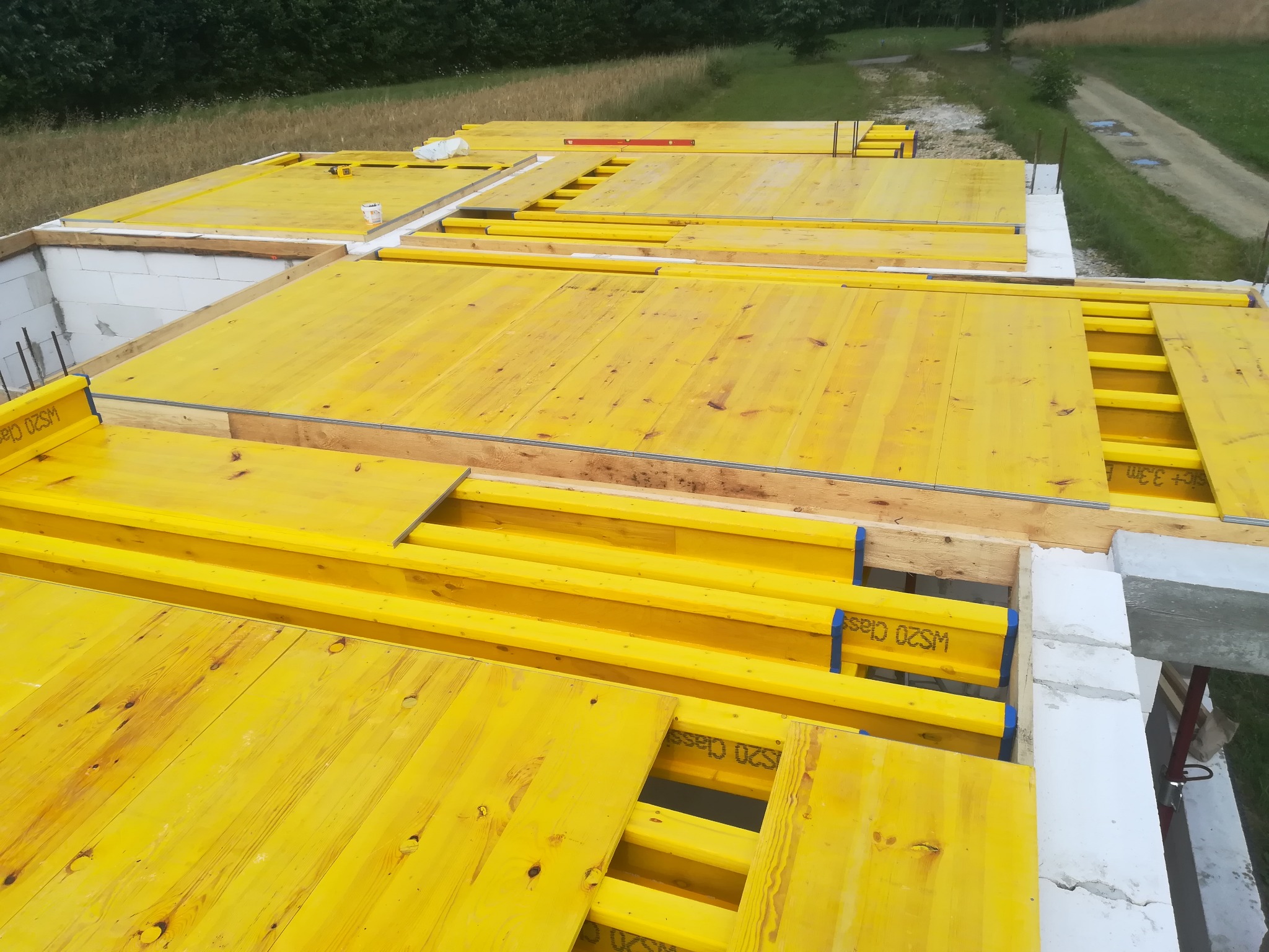 Wysoki kąt widzenia na szalunek stropowy z żółtych płyt OSB i belek WS20 Class, ułożony na białych bloczkach fundamentowych, widok na zielone otoczenie i drogę gruntową.