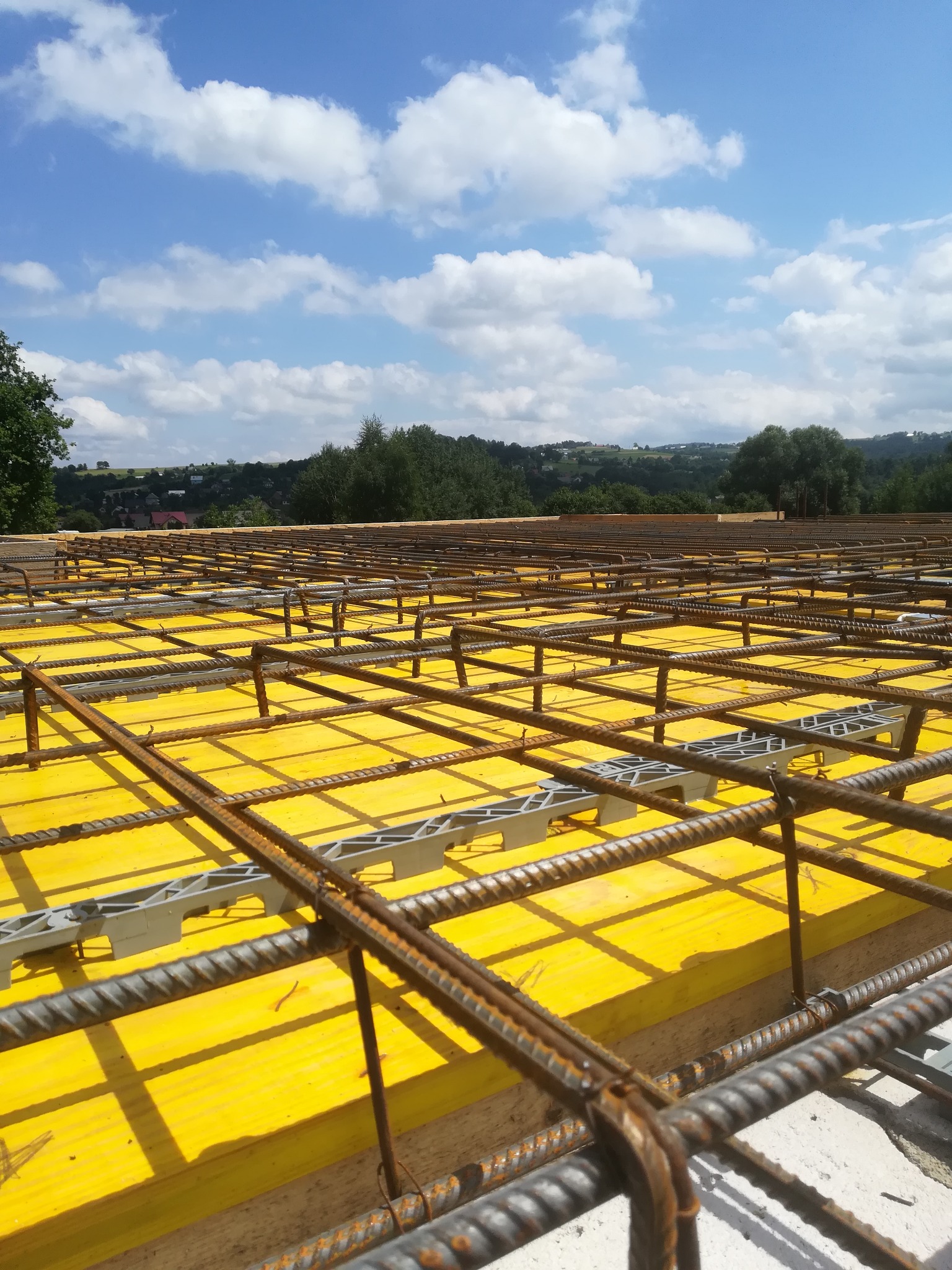 Żelbetowa konstrukcja stropu z żółtym szalunkiem i siatką zbrojeniową, widok z góry na tle krajobrazu Słopnic z błękitnym niebem.
