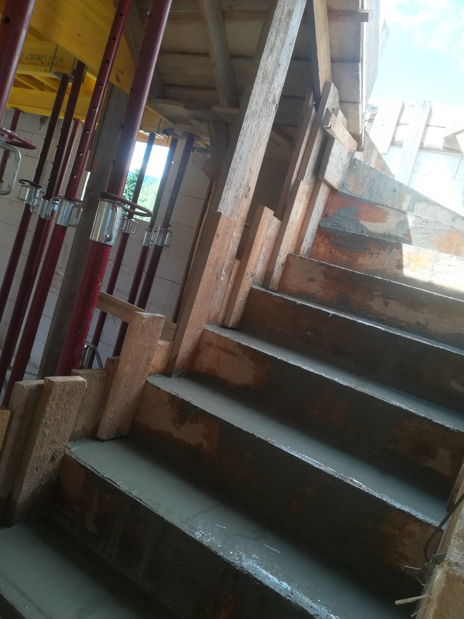 Wnętrze w trakcie budowy schodów żelbetowych, drewniane szalunki wypełnione świeżym betonem, widoczne podpory stemplowe i elementy konstrukcyjne budynku w tle.