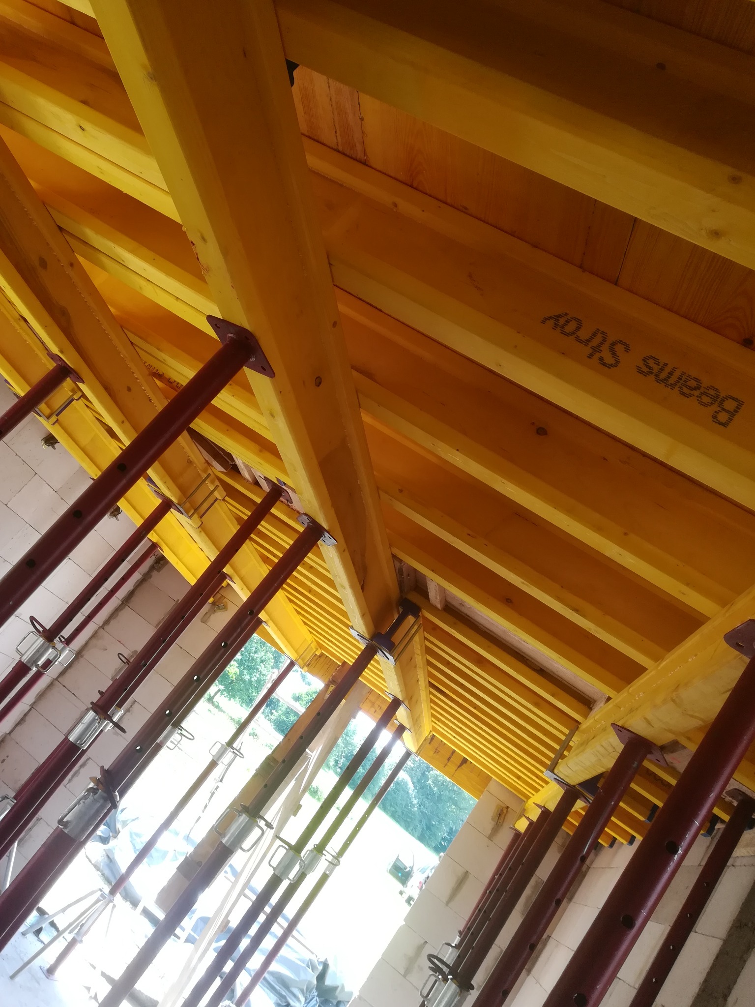 Żółte szalunki stropowe podparte czerwonymi stemplami teleskopowymi, widok z dołu na budowie domu z pustaków. Widoczny napis 'Beams Stroj'.