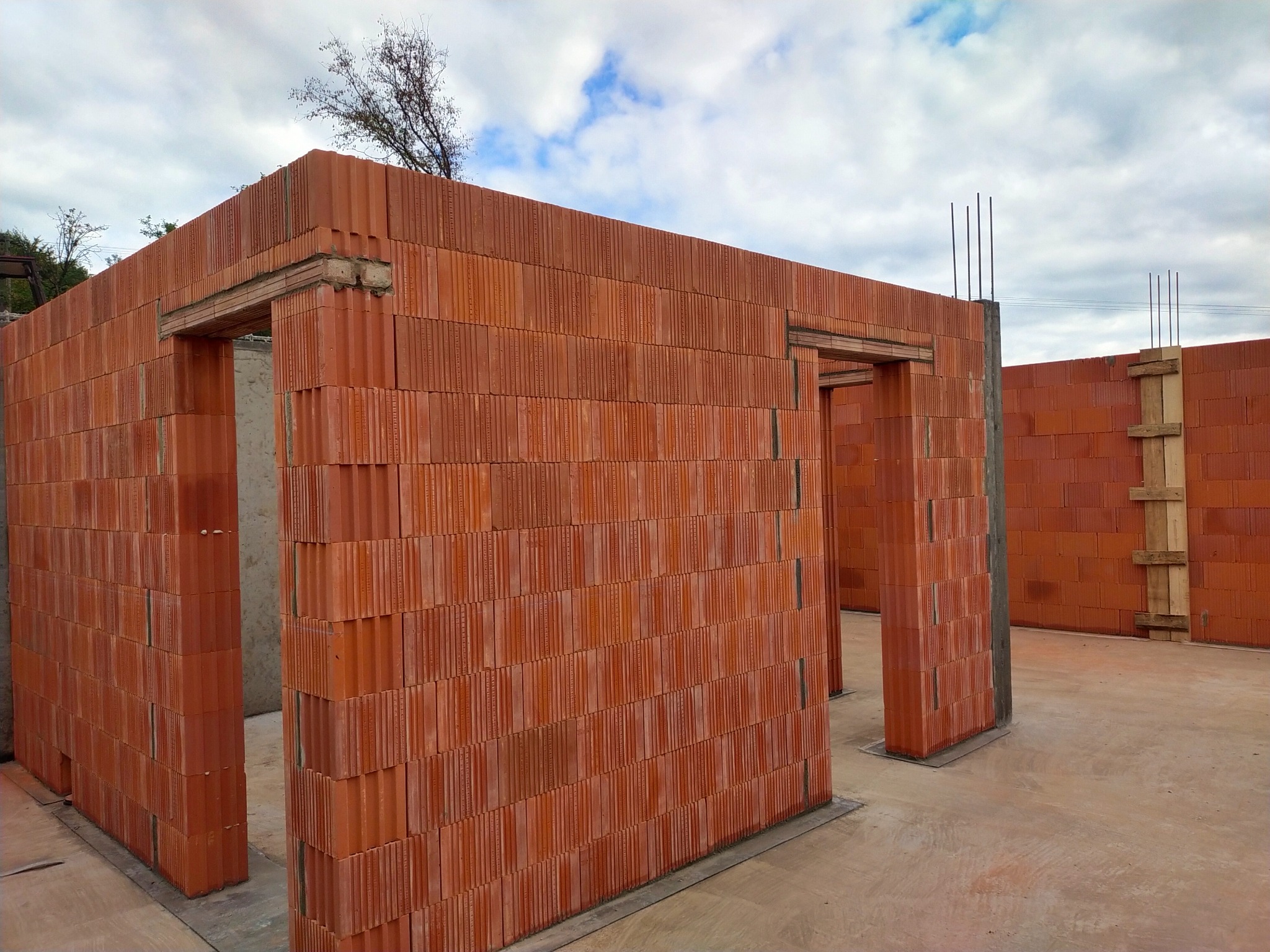 Wznoszenie ścian budynku z czerwonej ceramiki poryzowanej. Widoczne otwory drzwiowe z nadprożami i zbrojenie pionowe. Ściany na fundamencie, w tle pochmurne niebo.