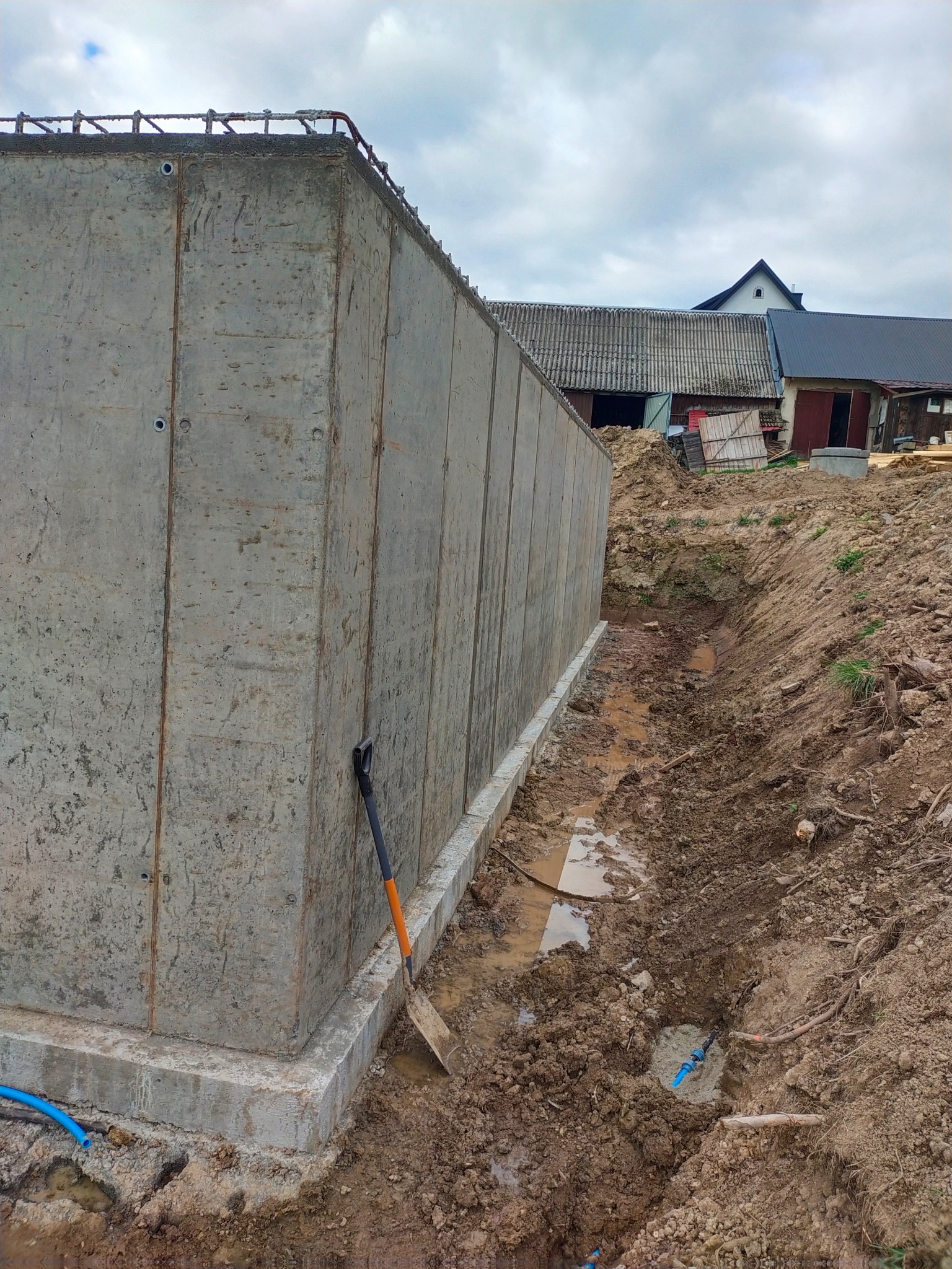 Betonowa ściana fundamentowa z wystającym zbrojeniem, widoczna łopata i instalacja. W tle budynki gospodarcze pod pochmurnym niebem.