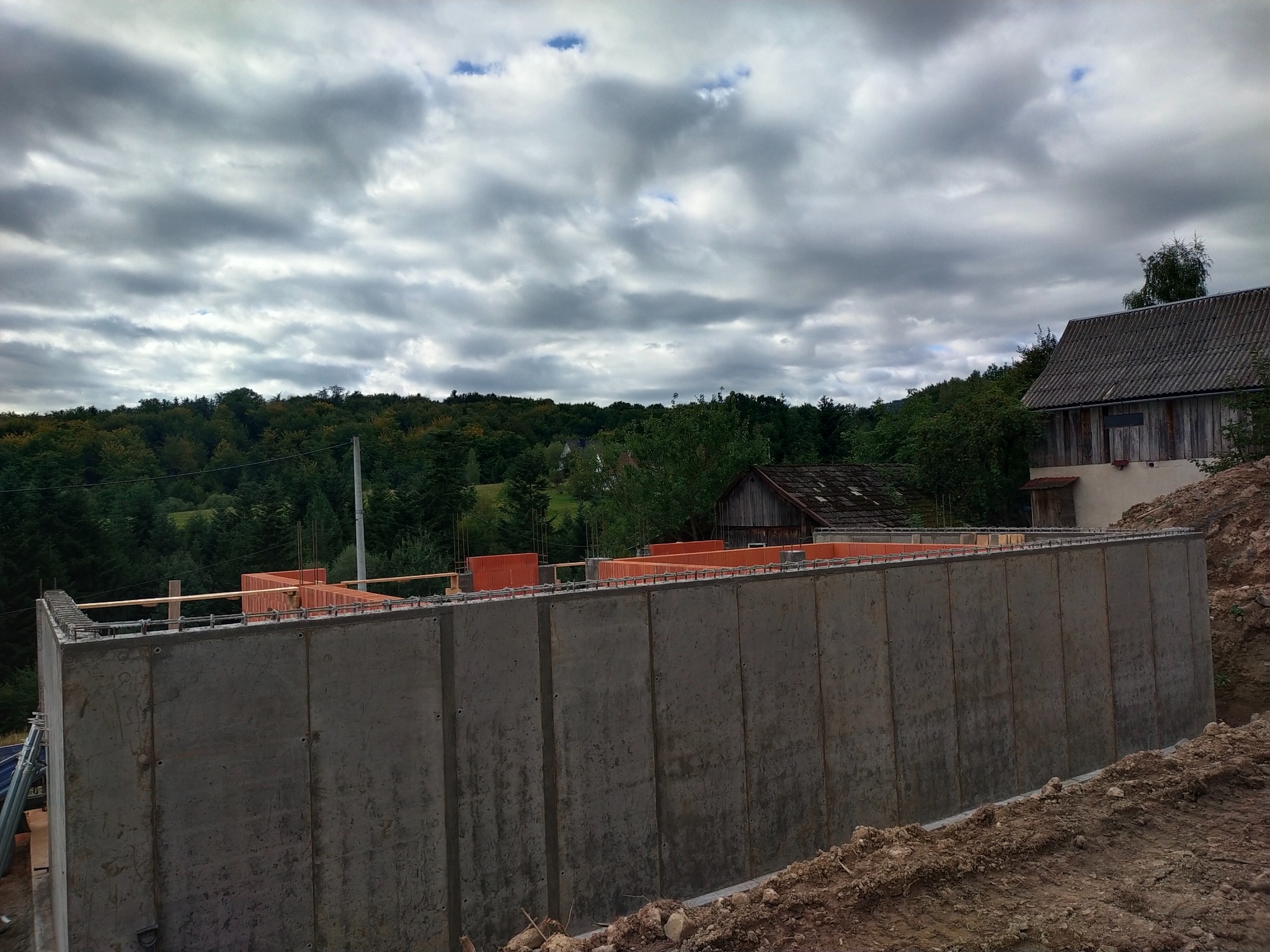 Betonowa ściana fundamentowa w trakcie budowy, widok na zalesione wzgórze i pochmurne niebo. Pomarańczowe elementy szalunku na górze ściany.