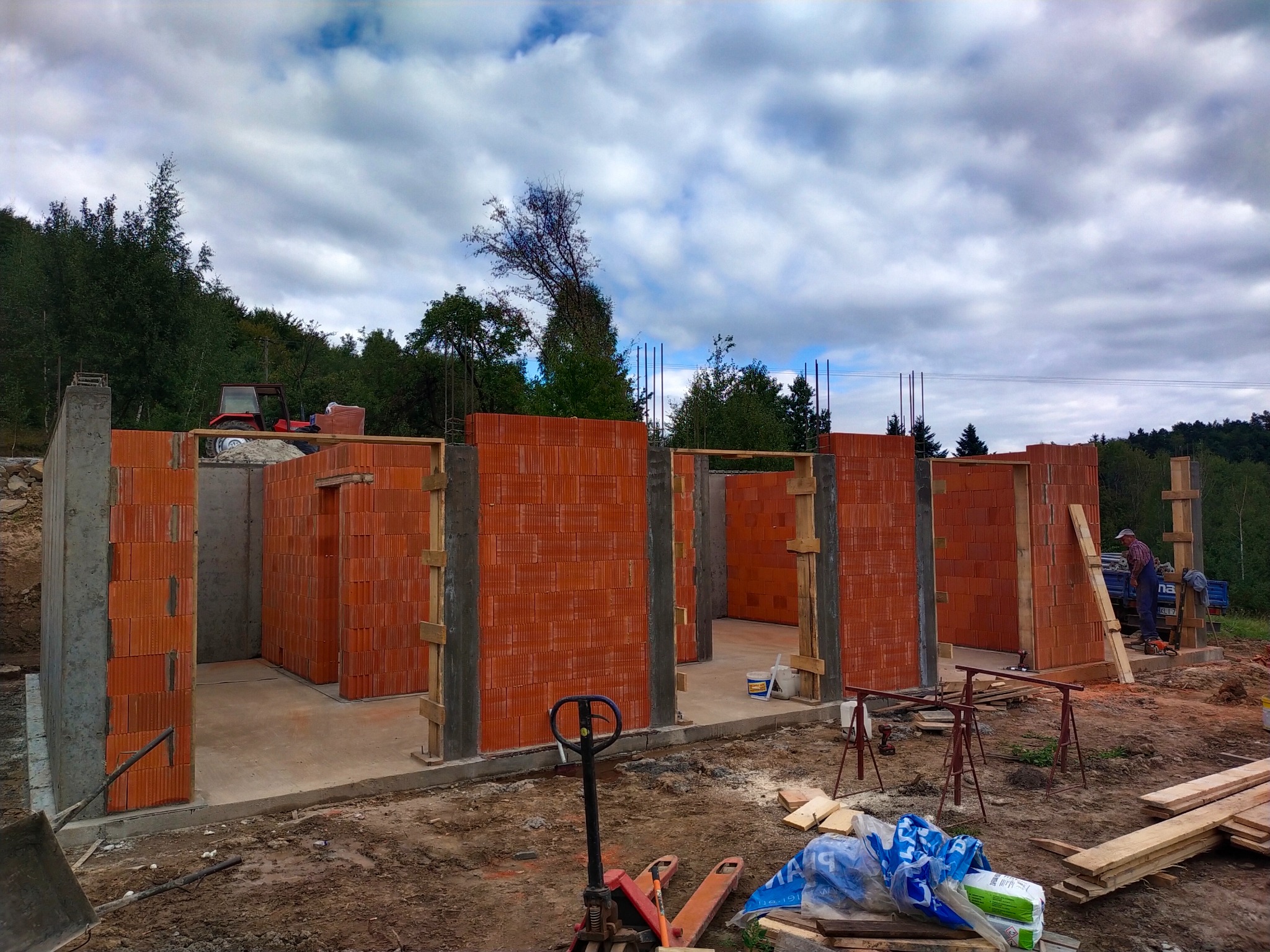 Wznoszenie ścian domu z czerwonej cegły. Widoczne betonowe słupy, drewniane szalunki i narzędzia. W tle zielone drzewa i pochmurne niebo.