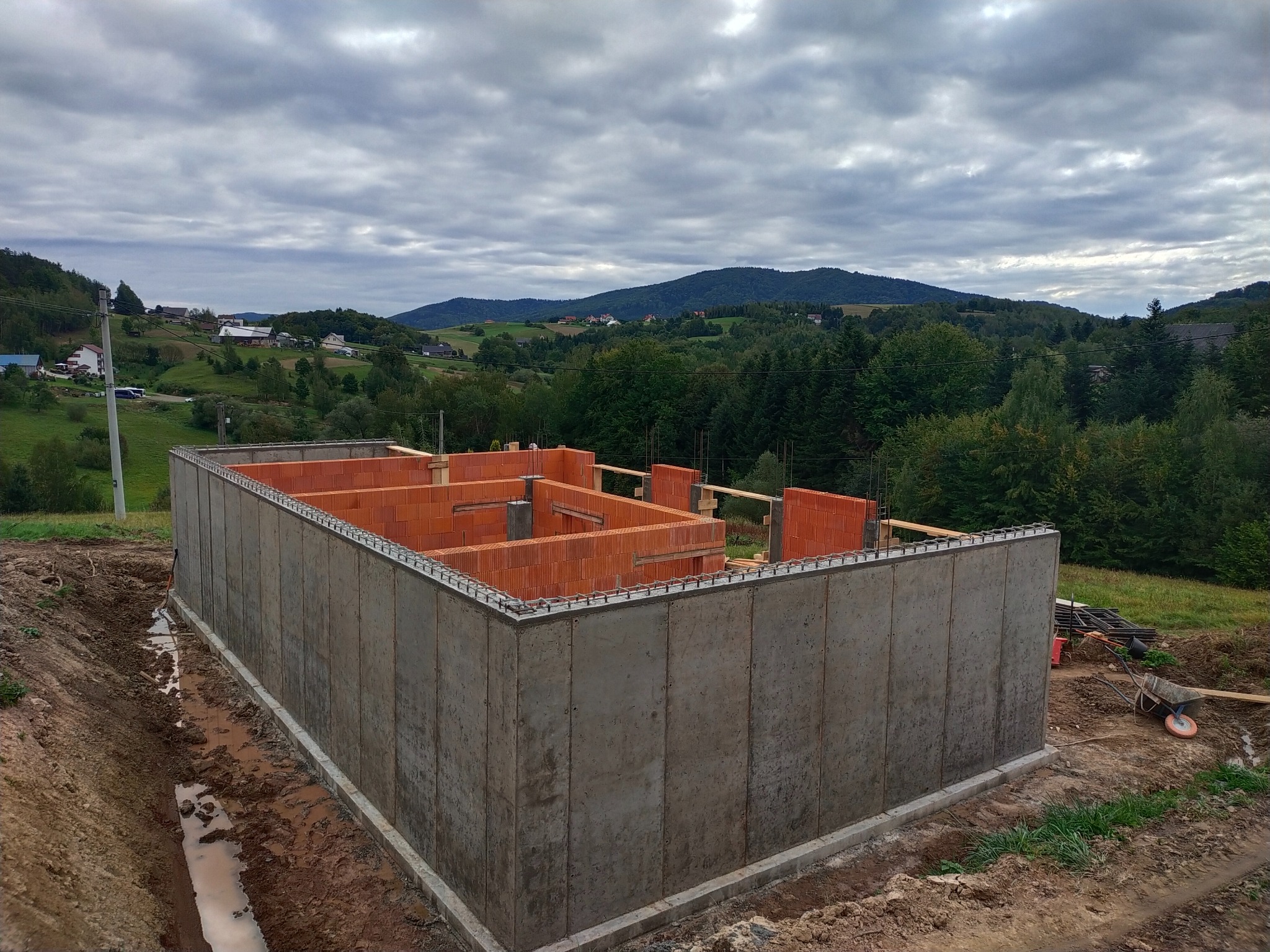 Surowe, betonowe fundamenty domu z częściowo wzniesionymi ścianami z cegły, na tle malowniczego, pagórkowatego krajobrazu pod pochmurnym niebem.