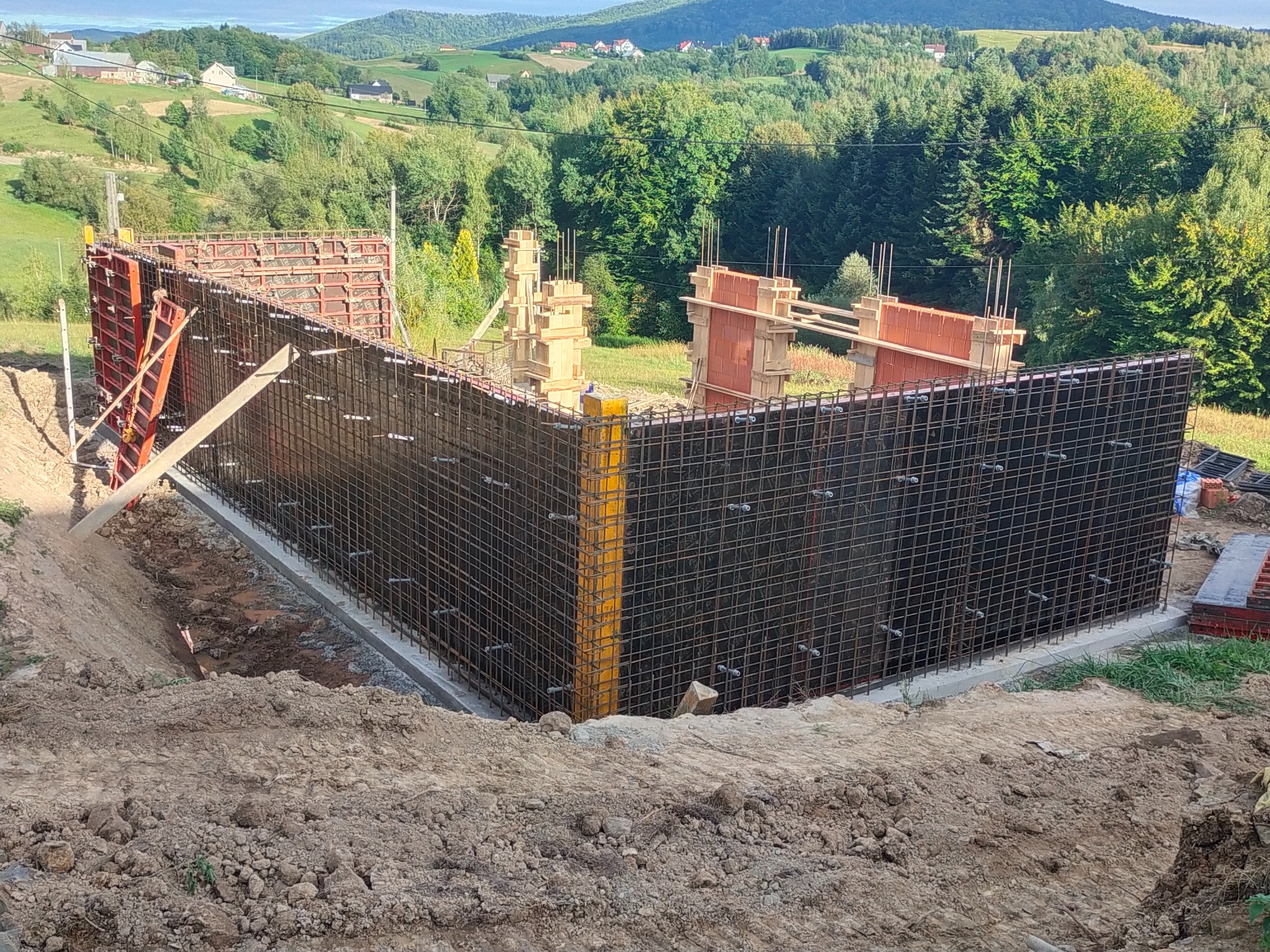 Zbrojenie i szalunek fundamentów pod budowę domu na tle krajobrazu. Widoczne metalowe pręty i drewniane podpory. Etap budowy z bliska.