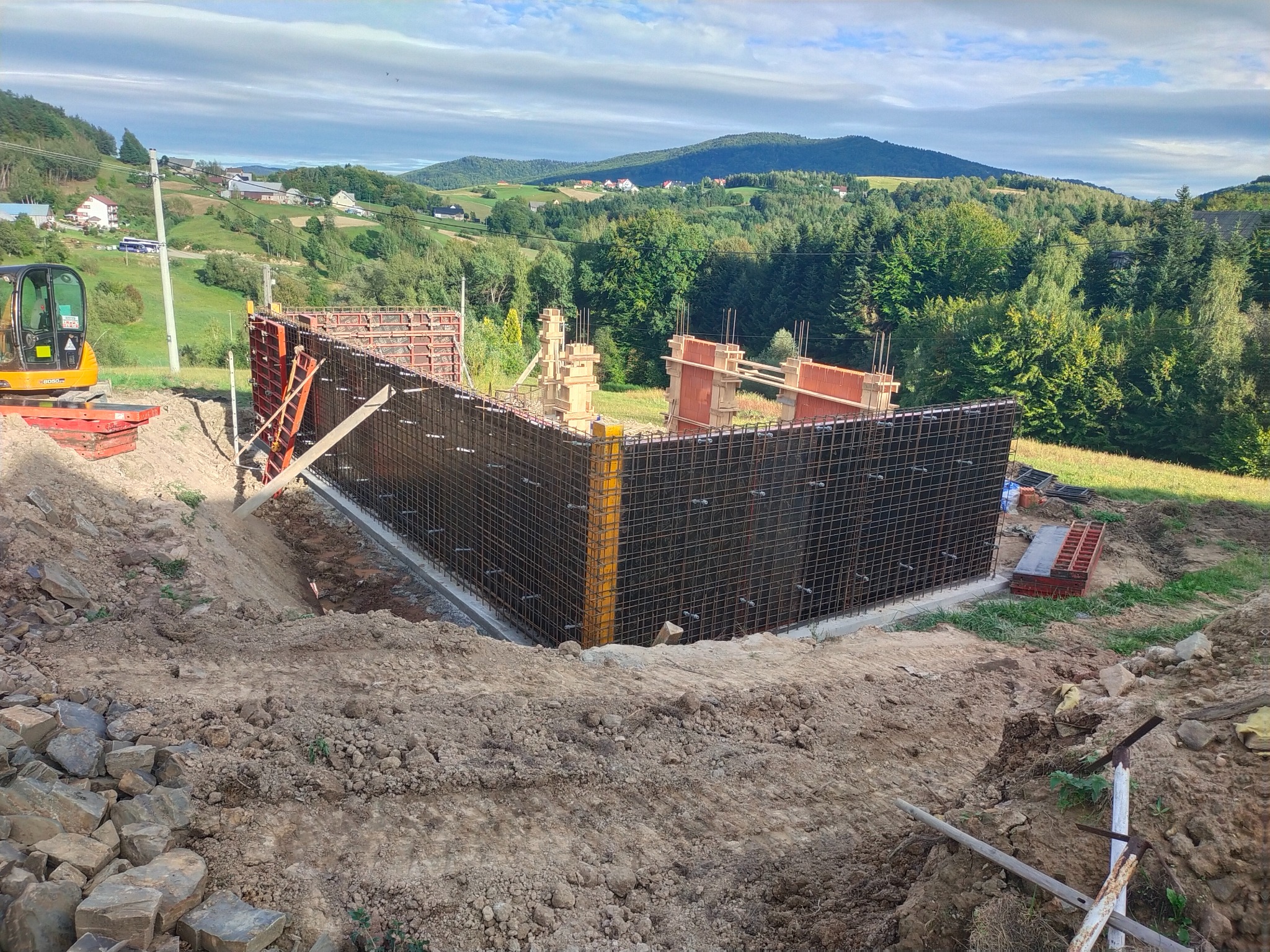 Zbrojenie fundamentów pod budowę domu na wzgórzu, widok na konstrukcję z prętów, szalunki i mini koparkę, malowniczy krajobraz w tle.